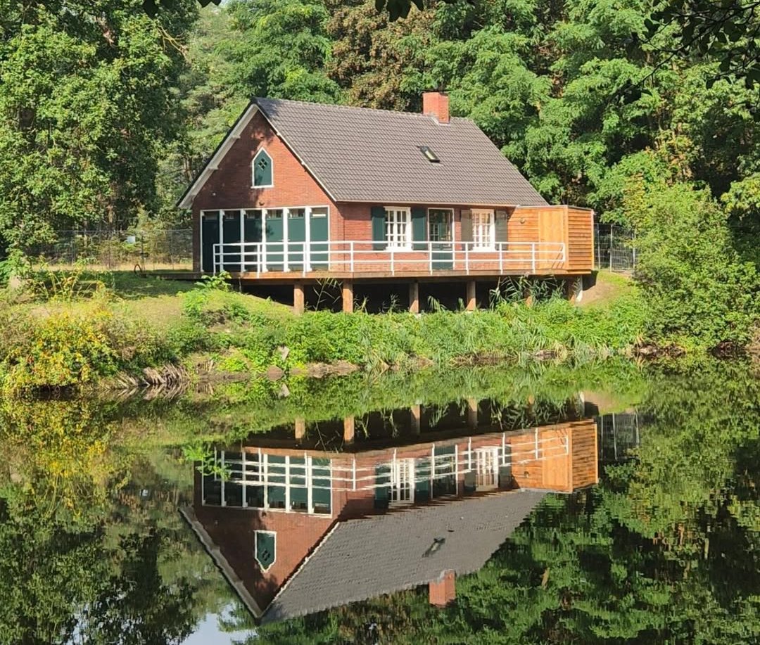 Het gerestaureerde Bakkershuisje van Coudewater, een rood bakstenen huisje met groene luiken en een houten veranda, weerspiegeld in de vijver tussen het groen